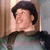 A person wearing a dark beret and green top with black shoulder accents, smiling while turning head left to right