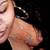 A close-up of a person with dark hair and large hoop earrings, wearing a light brown top with silver stripes, against a black background.