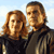 Static pixelated image of two people standing side-by-side against a light blue sky background with white clouds, rendered in low-resolution 256x256 pixels.
