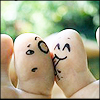 Two fingers with drawn faces kiss against a blurred green background, one with wide eyes and the other with closed eyes and a smile.