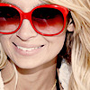 A close-up of a person with blonde hair wearing large red sunglasses and a white feathered earring, smiling with visible teeth.