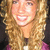 A woman with long curly blonde hair smiles broadly, wearing a purple top against a dark background.