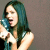 Pixelated image of a person with long teal hair holding a blue microphone, wearing a white sleeveless top against a gray background.