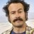 Static pixelated image of a person with dark hair and mustache, wearing a dark jacket over a blue shirt against a light background.