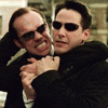 Two men in dark suits embrace; one wears sunglasses and holds a silver object near the other's face, with green text reading "THINK UNSLASHY THOUGHTS."