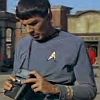 A person in a gray uniform with a white insignia holds a small device, standing before a blurred urban background.