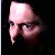Pixelated still image of a figure with a long nose, dark eyes, and a light-colored head covering against a dark background.