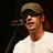 Pixelated image of a person wearing a cap and white shirt, standing before a microphone against a dark background with red text on the right.