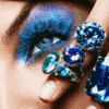 A close-up of a person's eye with blue eyeshadow and multiple blue gem-like objects near the nose and cheek, with a finger partially visible.