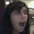 A pixelated close-up of a person with dark hair, mouth open in a shout or scream, against a blurred background with light and dark areas.