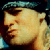 Pixelated close-up of a person with light skin and long hair wearing a dark cap, set against a dark background with blue and green accents.