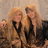 Two figures with long blonde hair stand side-by-side against a muted background, wearing dark clothing and holding hands.