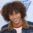 A pixelated image shows a person with curly brown hair wearing a dark jacket over a white shirt with red collar, next to a bright pink background with yellow text.