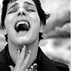 Black-and-white close-up of a person with short dark hair shouting with mouth wide open, hand near chin, against blurred background.