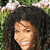 Pixelated close-up of a person with voluminous dark curly hair against green foliage background, smiling expression.