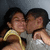 Two people in close embrace with one person kissing the other's cheek, wearing yellow and white clothing against a dark background.