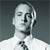 Pixelated grayscale portrait of a person with short hair, closed eyes, and a neutral expression, wearing a white shirt and dark tie against a gradient background.