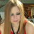 A pixelated image of a person with long light brown hair wearing a red top and dark necklace, set against a blurred background with indistinct shapes.