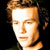 Pixelated static image of a person's face with light brown hair and neutral expression against dark background.