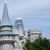 Pixelated static image shows a tall spire, multiple buildings, and green foliage under a light blue sky.