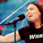 A person in a black shirt with partial white text stands before a microphone against a blue background with diagonal lines.