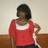 A person with short dark hair wears a red top with white trim and dark pants, standing against a light wall with a brown baseboard.