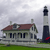 Pixelated static image of a two-story house with a red roof and a tall black-and-white lighthouse on a grassy area under a cloudy sky.