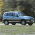 A light blue station wagon with dark windows and black wheels is positioned on a grassy area against a blurred background of trees and foliage.