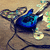Blue electric guitar with black neck and headstock, resting on brown textured surface with white circular accents, surrounded by abstract light patterns.