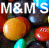 Pixelated static image of M&M's candy branding with large text and three colorful candies against a dark background.