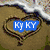 A heart-shaped sand drawing with a blue text overlay reading 'Ky KY' on the left, and a sunset over water with palm trees on the right.