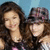 Two women with long brown hair pose together against a blue background; one wears a plaid hat and striped shirt, the other a pink top.