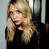 A static image of a person with long blonde hair wearing a black top and red lipstick, positioned against a light-colored wall and dark vertical structure.
