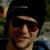 Pixelated static image of a person wearing dark sunglasses and a black cap, with visible facial features including nose and mouth, set against a blurred background.