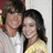 Two people in close physical proximity, one wearing a white shirt and the other in yellow, with dark hair on both figures against a light background.
