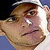 Pixelated close-up of a person's face with light brown hair, wearing a white cap and dark clothing against a blue background.