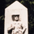 A static icon with a white house-shaped background, dark green foliage on the sides, and a central figure holding a small object.