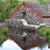 A diamond-shaped icon with a red border, featuring a gray stone house with a dark door and white trim, set against a green and gray background.
