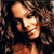 Pixelated close-up of a person with curly brown hair, light skin, and a dark top, set against a dark background with brown and black tones.