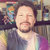 Pixelated static image of a person with closed eyes, light skin, and a goatee, wearing a dark jacket over a light shirt, set against a blurred background with colorful objects.