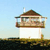 Pixelated image of a small house with a brown roof, white walls, and a balcony, set against a light blue sky and dark ground.