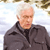 Pixelated image of a person with short white hair wearing a dark gray zip-up jacket over a black shirt, standing against a light blue and white background.