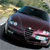 A dark-colored sedan with visible headlights and grille, positioned on a road with blurred background elements suggesting motion or speed.