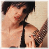 A person with short dark hair and red lipstick holds a guitar, wearing a black sleeveless top against a light background.