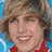 Pixelated image of a smiling person with light brown hair wearing a red top against a blue background, next to red pixelated text on black.