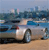 Pixelated static image of a light-colored car on a sandy beach with blue water and distant buildings under a pale sky.