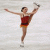 Pixelated static image of a figure skater in mid-pose with arms extended and one leg lifted, wearing red top and orange shorts against light background.