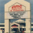 Pixelated static image of a building facade with a red sign above a dark entrance, set against a light background.