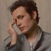 A person with dark curly hair wearing a light gray collared shirt, resting their right hand on their forehead against a plain beige background.