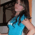 A person in a blue sleeveless top with long dark hair, standing against an indoor background with wooden railings and light-colored walls.